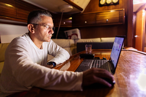 Mature man working on laptop on sailing and sitting in sailboat salon. He has wireless headphones in ears and wearing eyeglasses.