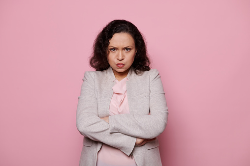 Angry dissatisfied middle-aged multiethnic woman in a light gray jacket, get bored upset, expressing worry anxiety, posing with lips pouted and arms folded on pink background. People Emotions Concepts