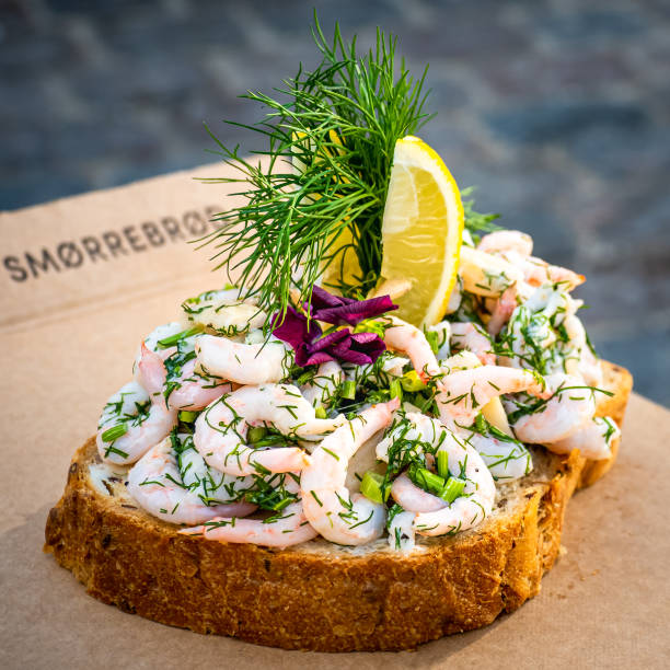 Close-up of a traditional Danish delicacy sandwich, adorned with prawns, dill, chives, lemon slices, and edible flowers, with the lettering smørrebrød, engl. butter and bread, in the background. Traditional delicious Danish snack called smørrebrød butter bread, rich adorned with prawns, chives, dill, lemon slices and edible flowers, showcasing the delicacy of Scandinavian food and culture. danish culture stock pictures, royalty-free photos & images