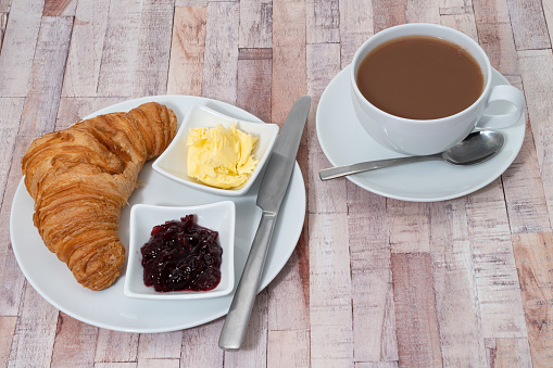 Breakfast items of a fresh croissant with dairy butter and raspberry jam and a cup of tea