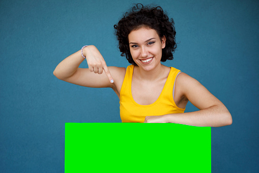 Young woman pointing at blank green advertising board against blue background