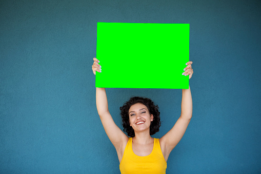 Young woman holding blank green sign board against blue background