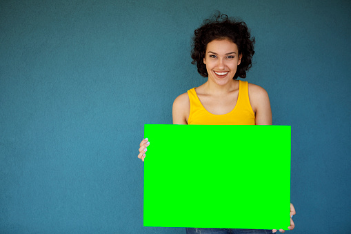Young woman holding blank green sign board against blue background