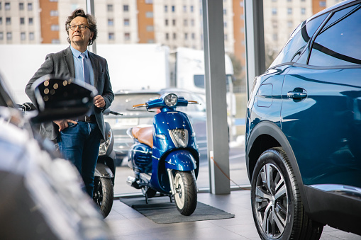 Businessman with short brown hair and glasses,wearing a suit,is standing next to a brand new car at his car dealership,looking at a customer in the distance with serious facial expression,scooters in the back