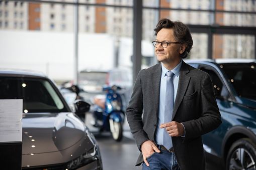 Male adult car seller with short brown hair and glasses,wearing a suit,is waiting for a customer,standing indoors at his car dealership with one hand in his back,brand new cars and scooters in the background