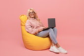 Happy freelancer lady using laptop, sitting in beanbag chair and smiling, checking new website on pink background