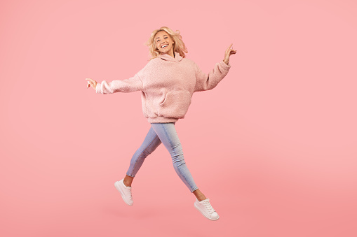 Joyful stylish woman running and jumping, posing in mid-air over pink studio background, copy space. Joy and happiness, positive emotions concept. Full length shot