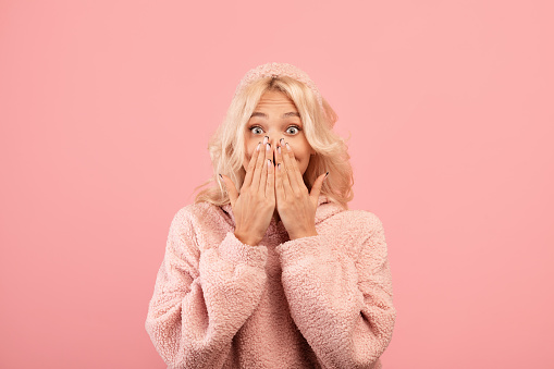 Human emotions concept. Shocked lady covering her mouth with hands and looking at camera, standing isolated over pink studio background