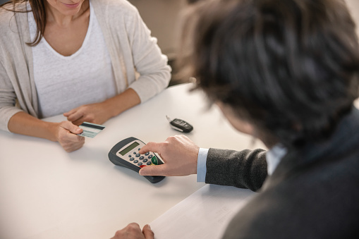 Car salesman with short brown hair is typing the amount the customer has to pay for a brand new car in his credit card reader,sitting together with a female customer with long brown hair at his office table indoors at a car dealer,woman holding her credit card,car keys lying next to her on the table