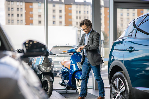 Adult car dealer businessman with short brown hair and glasses,wearing a suit,is looking down at his clock,walking indoors at his car dealership with serious facial expression,brand new cars in front,scooters in the back
