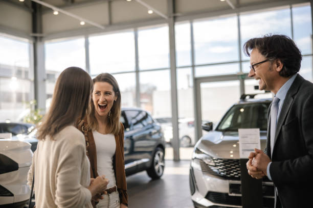 istock Two female customer looking at each other with amazed facial expression after hearing about an amazing deal from a male car seller,man standing next to them smiling,indoors at a car dealer 1472178902