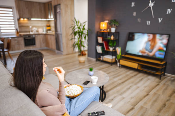 istock Young woman watching television and eating popcorn while sitting on a sofa at home 1472179071