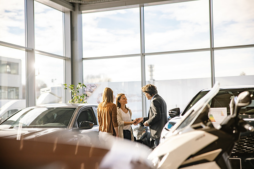 Businessman with short brown hair and glasses is giving the keys to a female customer with long brown hair,both smiling at each other,woman with blond hair standing in the front,indoors at a car dealership in the evening