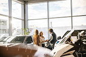 Businessman is giving the keys to a female customer,both smiling at each other,woman with standing in the front,indoors at a car dealership in the evening