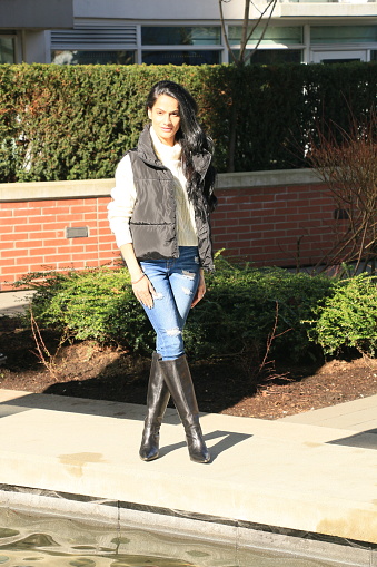 An Indian woman outside an apartment on a sunny day. She is wearing long, black, wavy hair, makeup, a white sweater with a gray vest, torn blue jeans, black knee length high heel boots. She is standing on a concrete walkway next to a pond and garden near apartments.