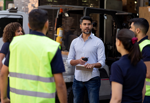 Latin American business manager talking to a group of workers at a distribution warehouse and coordinating a cargo delivery
