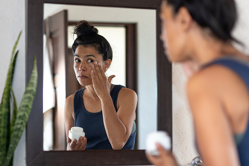 Woman applying facial moisturizing cream to face, looking into bathroom mirror