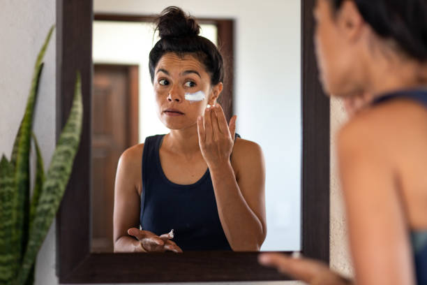 Woman applying white sunblock to face, looking at
 bathroom mirror stock photo