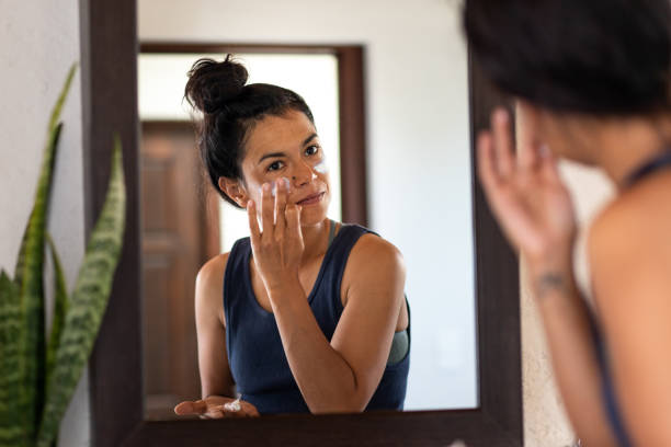 Donna che applica la protezione solare bianca al viso, guardando
 specchio da bagno - foto stock