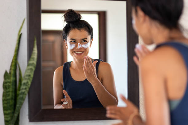 Woman applying white sunblock to face, looking at
 bathroom mirror, smiling stock photo