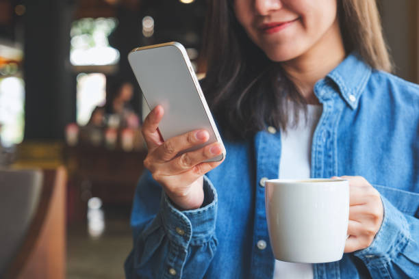 Closeup image of a young woman holding and using mobile phone while drinking coffee in cafe