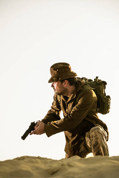 istock Soldier with pistol kneels on dune looking sideways 1472212329