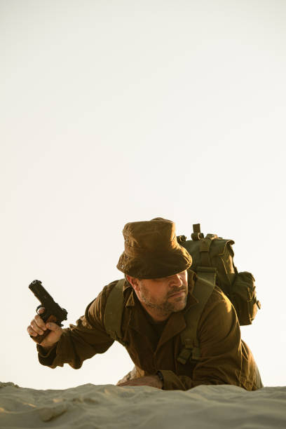 A soldier in uniform, alert to danger, holding a pistol crawling over a dune. Low angle view, portrait format for book cover purposes. Toned, copy space above.