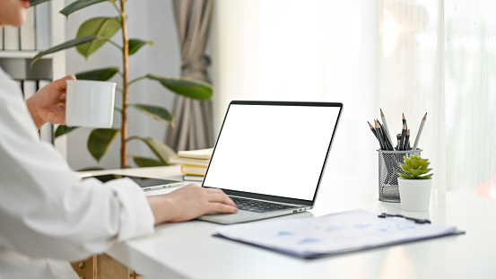 Cropped image of professional Asian businesswoman or female CEO sipping her morning coffee and using laptop at her office desk. laptop white screen mockup