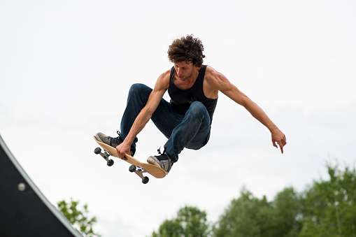 Athletic young man with curly hair wearing jeans and a sleeveless shirt is flying through the air after launching off a skateboard ramp in a park. He is holding his skateboard with one hand to his feet, paused in the air.