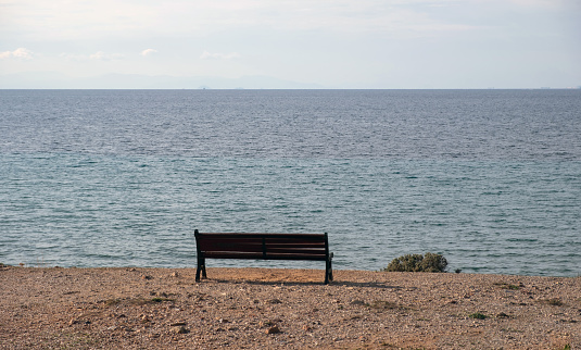 Lonely empty old wooden bench on soil over the beach. Winter sunny day, calm blue sea, cloudy sky background. Greek seascape.