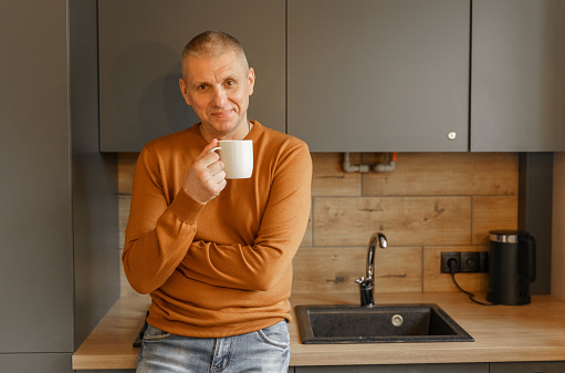 Portrait of a mature man in an orange jumper in the kitchen with a mug of warm tea.