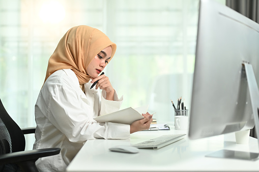 Focused millennial muslim businesswoman wearing hijab sitting at her workplace and checking working schedule plan.