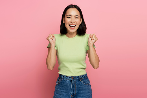 Portrait of excited overjoyed woman standing with raised fists and shouting yeah, I'm winner, rejoicing victory, success. Studio shot isolated on pink background