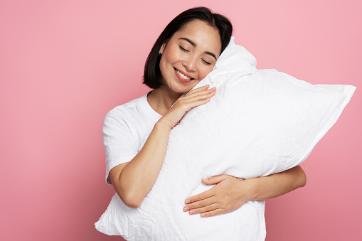 Satisfied gorgeous girl hugging pillow with closed eyes and sleeping isolated on pink coloured background. Bedtime concept