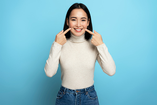 Portrait of pretty cheerful woman points index fingers at smile shows white teeth, looking at camera. Indoor studio shot isolated on blue background