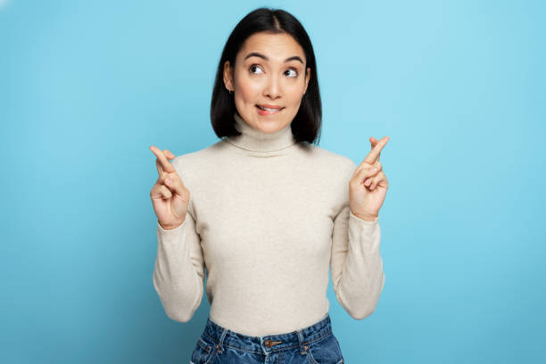 Hopeful nervous brown haired woman of young age crossing fingers for luck Hopeful nervous brown haired woman of young age crossing fingers for luck, hope for better, ritual, looks up with hopeful look. Indoor studio shot isolated on blue background fingers crossed stock pictures, royalty-free photos & images