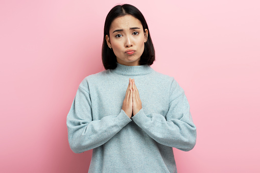 Portrait of attractive woman praying, begging with pleading grimace, gesture of asking apologising. Indoor studio shot isolated on pink background
