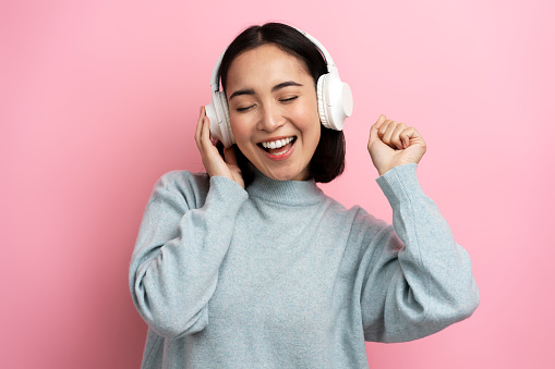 Happy satisfied girl standing listening favourite music with headphones, closed eyes and toothy smiling, Indoor studio shot, isolated on pink background