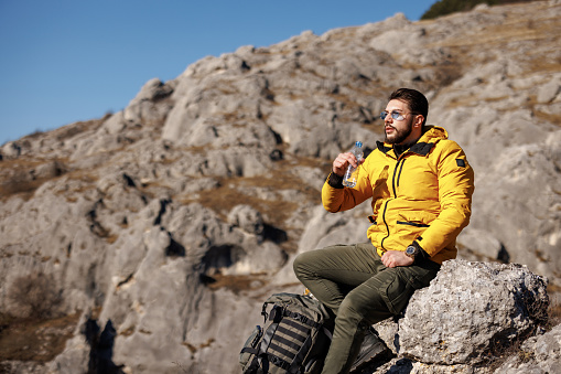 Young man sitting and drinking water while resting from hiking on rocky mountain alone
