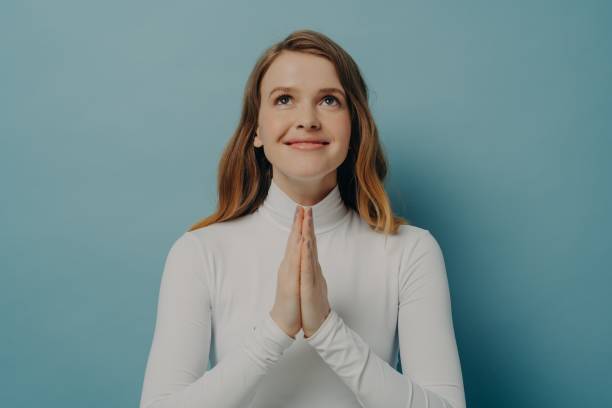 istock Happy young woman making wish while folded hands in prayer gesture, isolated on blue background 1472245425