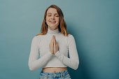 Smiling young woman holding hands folded in prayer gesture, keeping eyes closed in hope