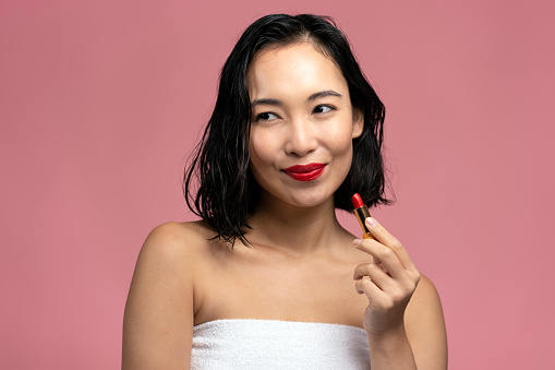 Young beautiful girl painting lips with lipstick. Asian woman applies makeup to her face with red smear. Beauty treatments concept