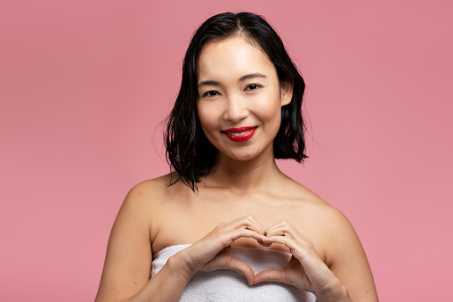 Young asian woman with bare shoulders showing heart shape with hands and looking camera isolated on pink background
