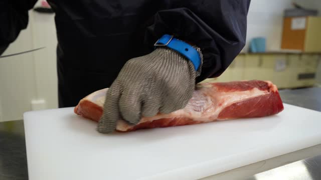 A Butcher in metal protective gloves Fills a piece of Pork Meat on a cutting white board with a Knife. Production line