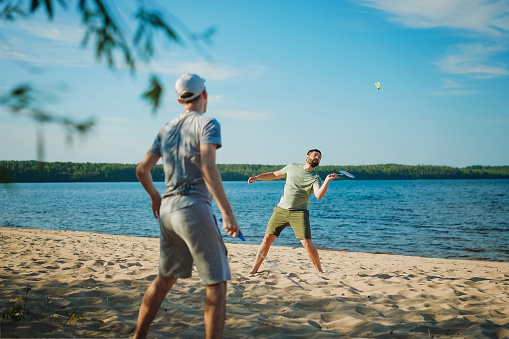 caucasian man and teenager son playing badminton on the lakeside. Parents and teens concept. Image with selective focus.