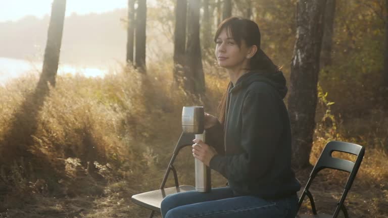 female sits on chair near campfire and drinks drink from thermos while enjoying