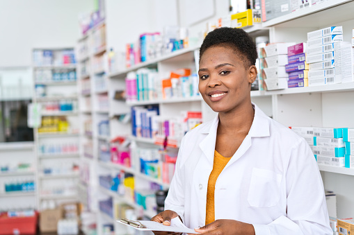 A young pharmacist standing in front of medicine selves, holding prescription in hands and smiling for the camera.