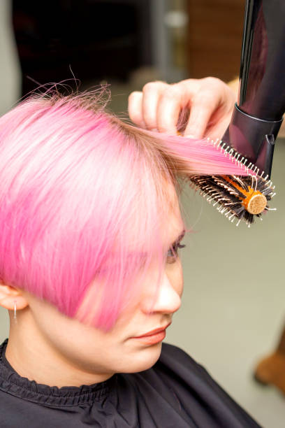 若い白人女性の短いピンクの髪を黒いドライヤーと黒い丸いブラシで、美容院の男性美容師の手で乾かし、接写する。 - frizzy human hair hairdresser hair dryer ストックフォトと画像