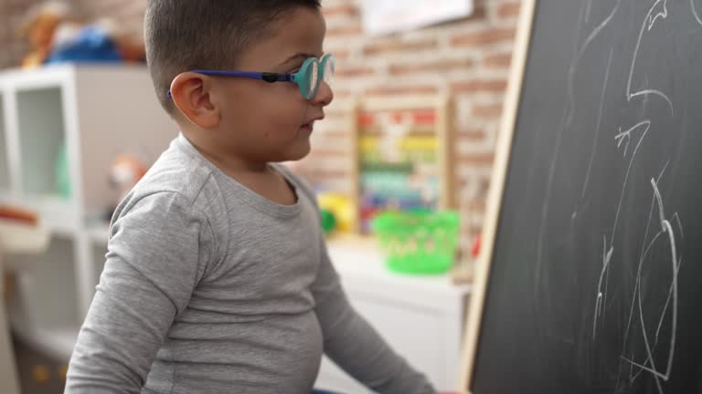 Adorable hispanic toddler student smiling confident drawing on blackboard at kindergarten