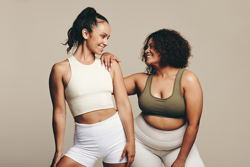 Female athletes of varying body shapes smile as they stand together, dressed in workout gear. Two confident young women showing their dedication to fitness and a healthy lifestyle.